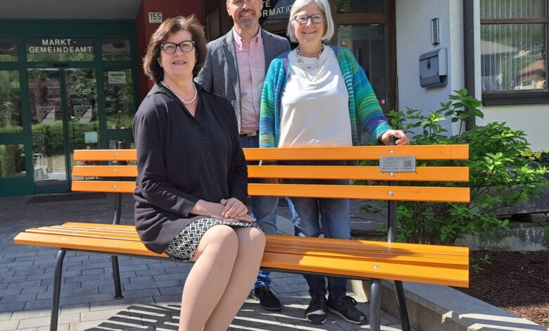 Frau sitzt auf oranger Bank, der Bürgermeister der Gemeinde Kirchbach und eine weitere Frau stehen dahinter. (c) Gemeinde Kirchbach